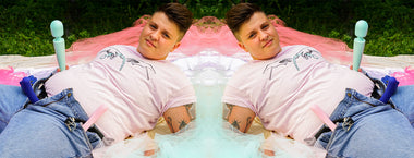 Photo of June Pilote lying down, holding wand vibrator and wearing strap on with dildo attached